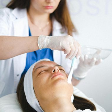 Concentrated female cosmetologist in medical uniform and gloves using brush to apply cosmetic mask on face of client with closed eyes