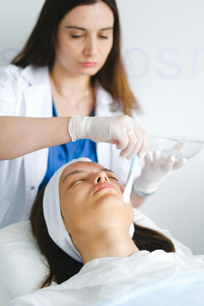 Concentrated female cosmetologist in medical uniform and gloves using brush to apply cosmetic mask on face of client with closed eyes